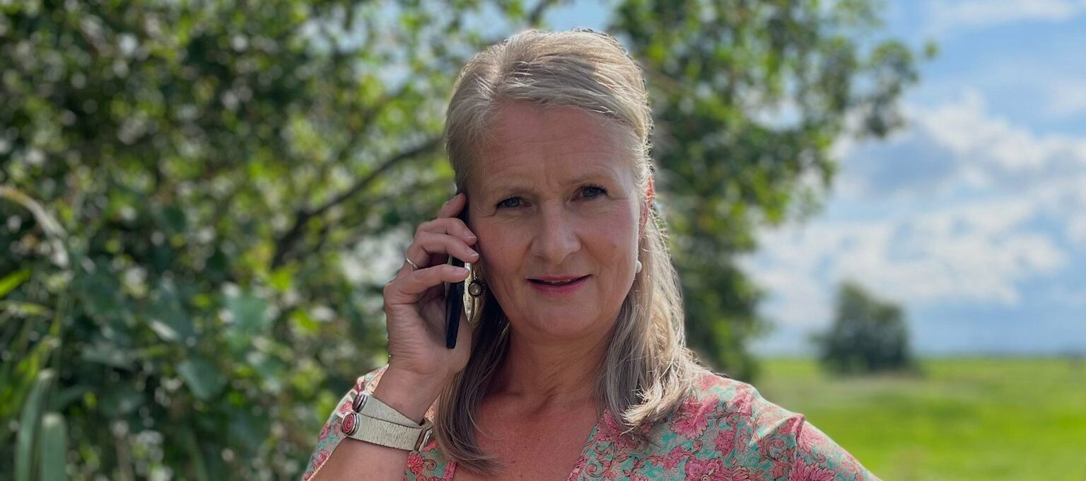 Mirjam staat op brug aan de telefoon. Achtergrond boom, weiland en blauwe lucht met wolkjes