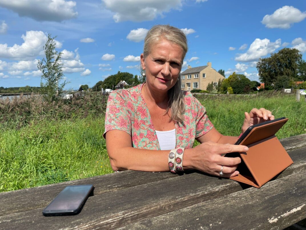 Mirjam aan tafel in park met laptop en telefoon. Achtergrond groen en IJsseldijk