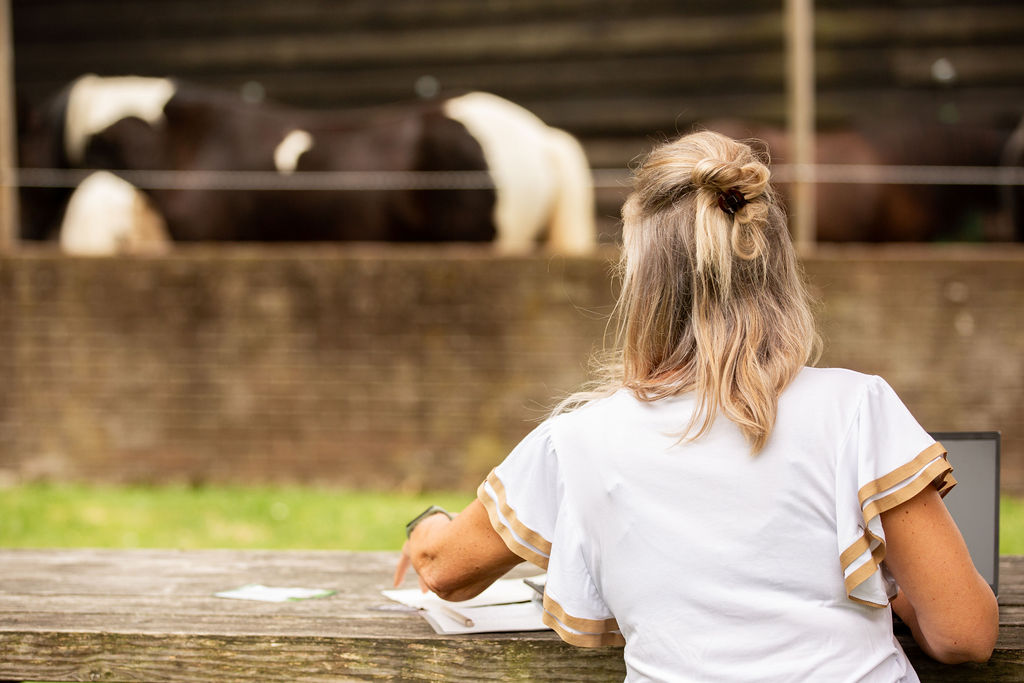 Mirjam aan het werk gezien vanaf de achterkant met paarden op de achtergrond